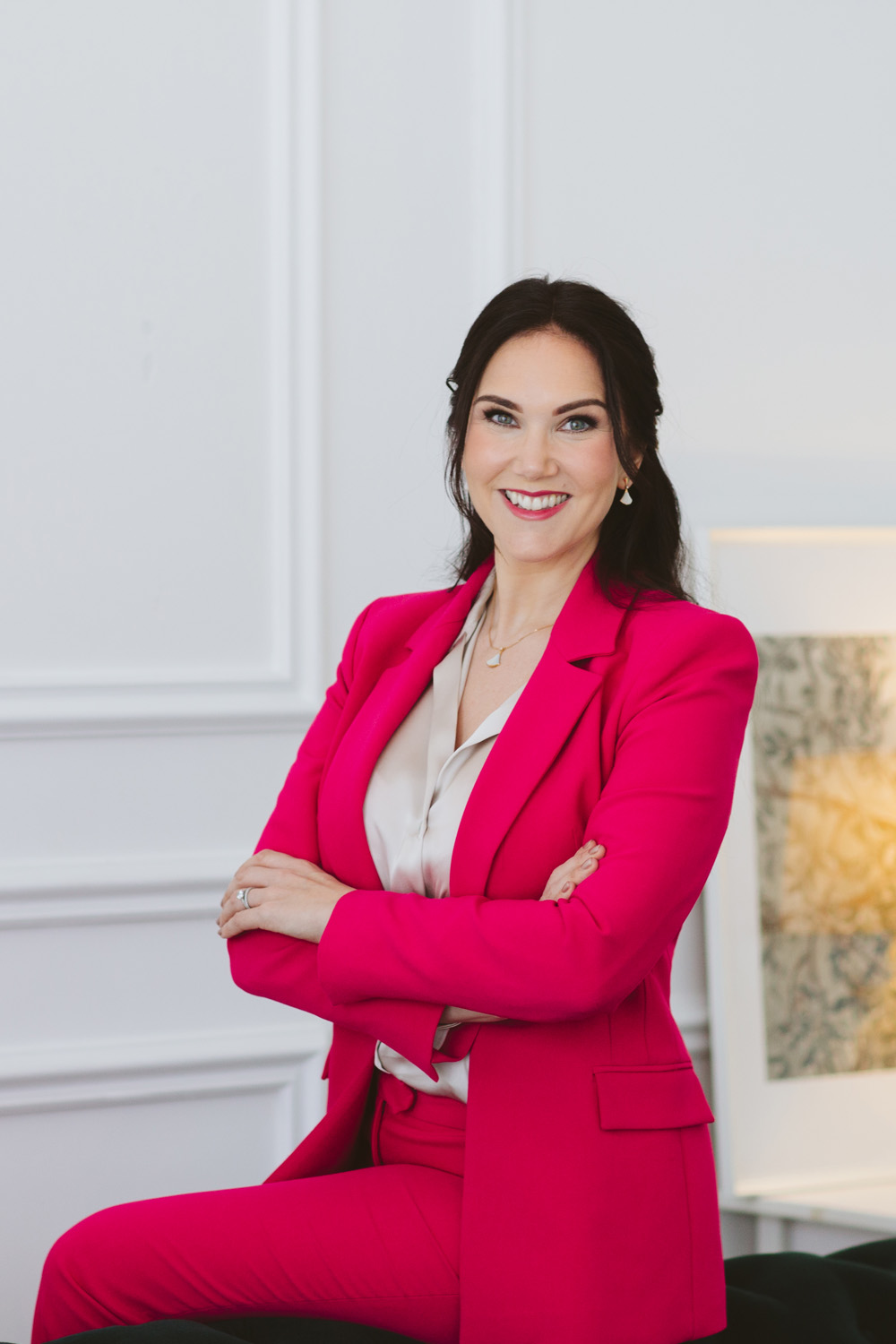 Victoria in a red suit sat on the arm of a sofa with arms crossed